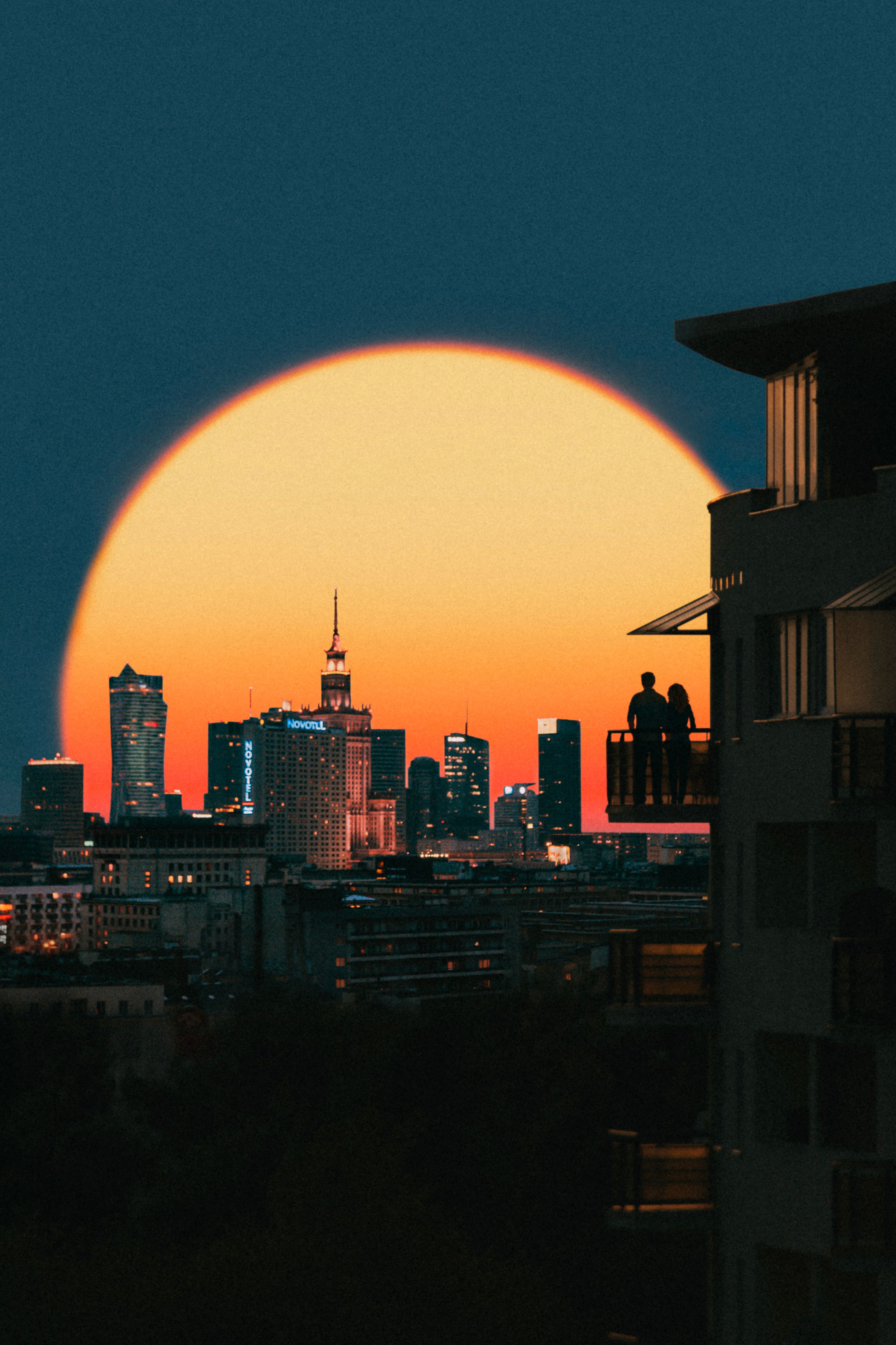Silhouetted couple on a balcony watching a dramatic sunset over a city skyline.