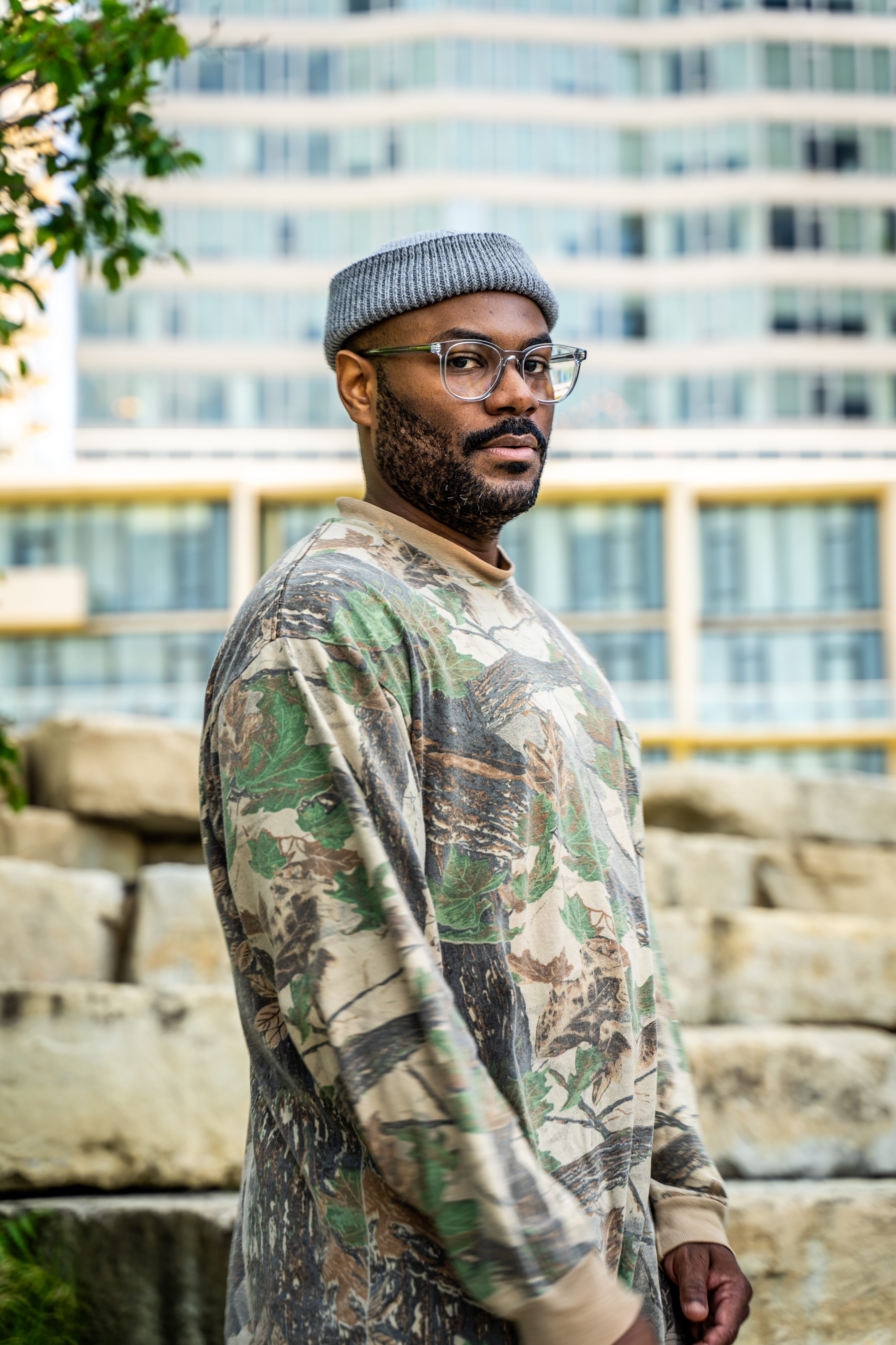Stylish man in camouflage sweater poses confidently in an urban outdoor setting with modern buildings in the background.