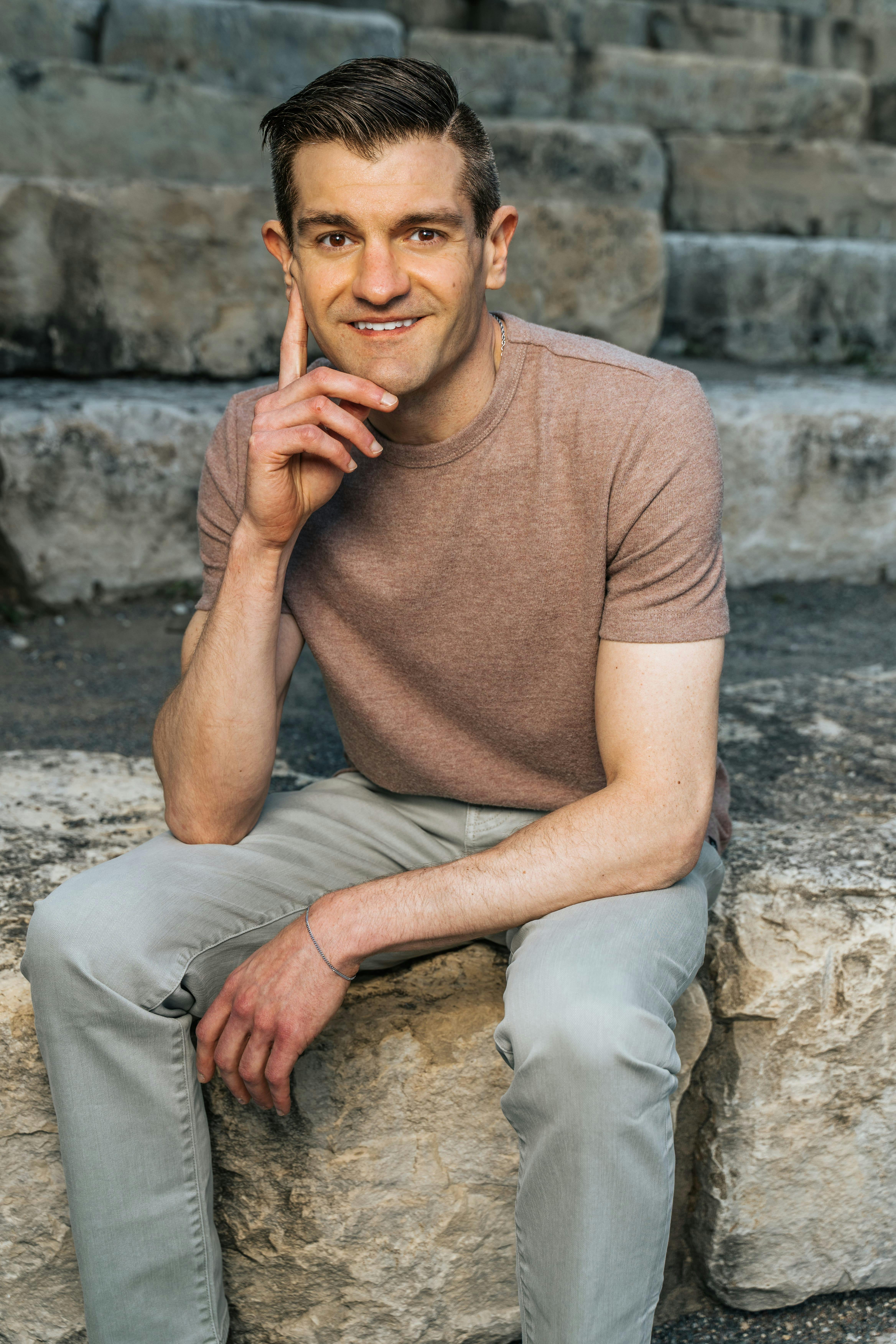 Confident man sitting casually on outdoor stone steps, smiling warmly at the camera.