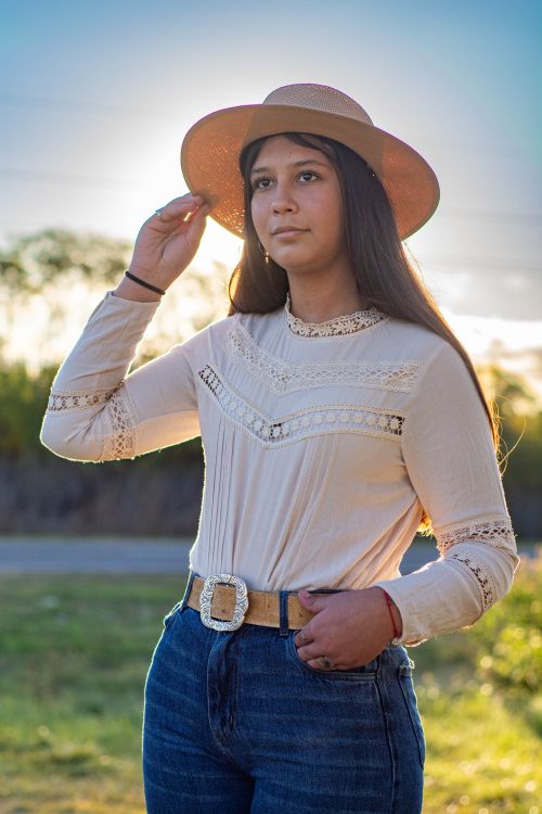 Free A young woman in casual attire stands outdoors with the sun setting behind her, wearing a stylish hat. Stock Photo