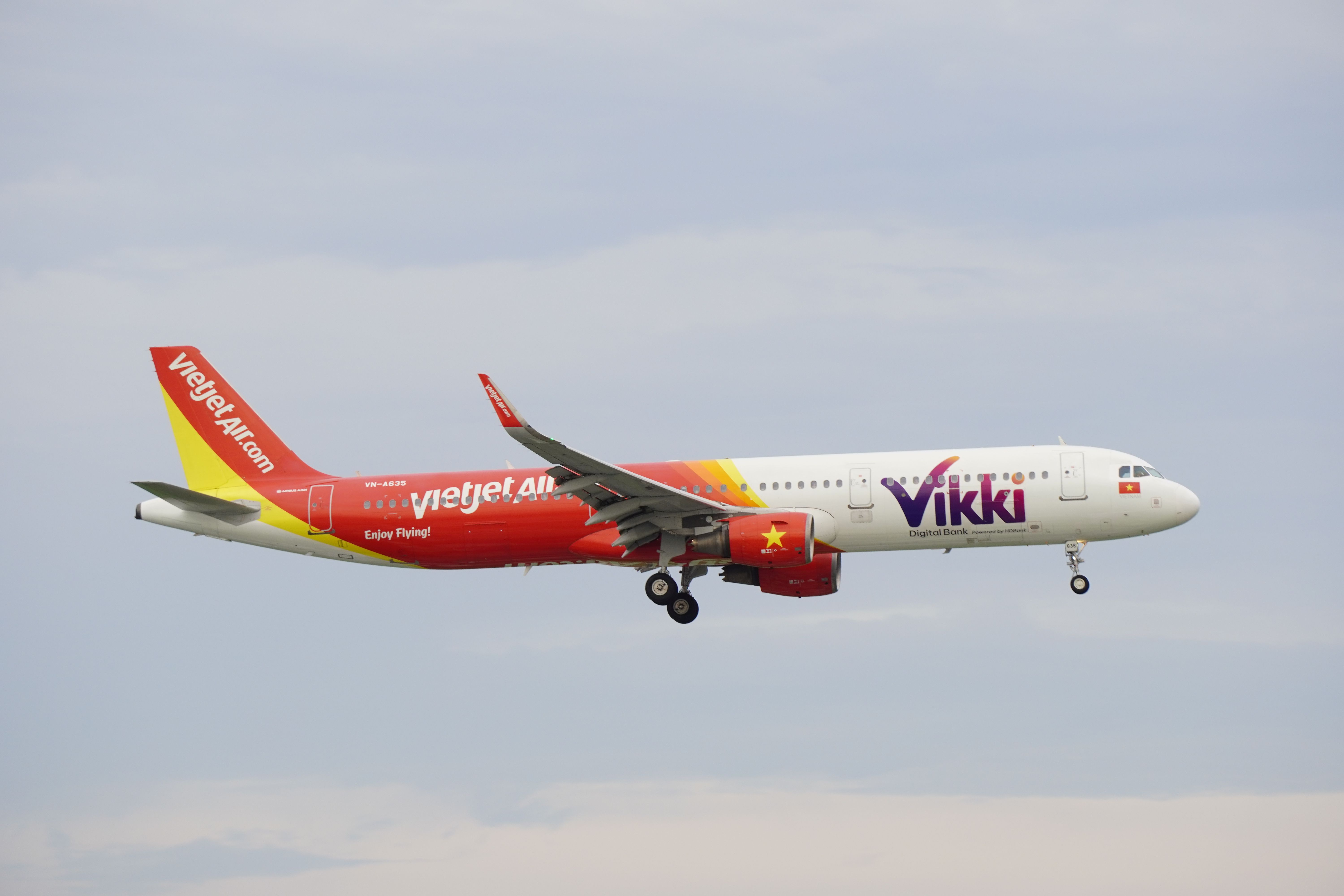 A VietJet airplane with Vikki branding in mid-air against a cloudy sky.