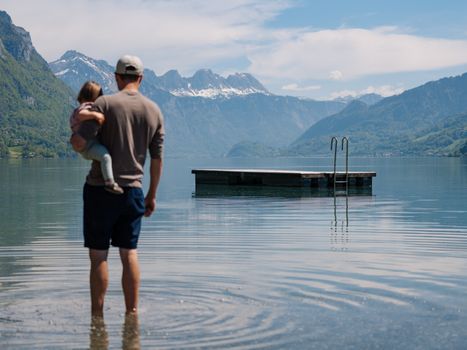 Father holding child while standing in a lake with a scenic mountain backdrop in Switzerland.