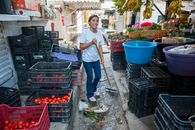 A woman cleans the aisle of an indoor produce market surrounded by fruits and vegetables.