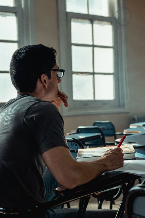 Free Male student in a classroom writing notes while studying alone. Quiet academic environment. Stock Photo