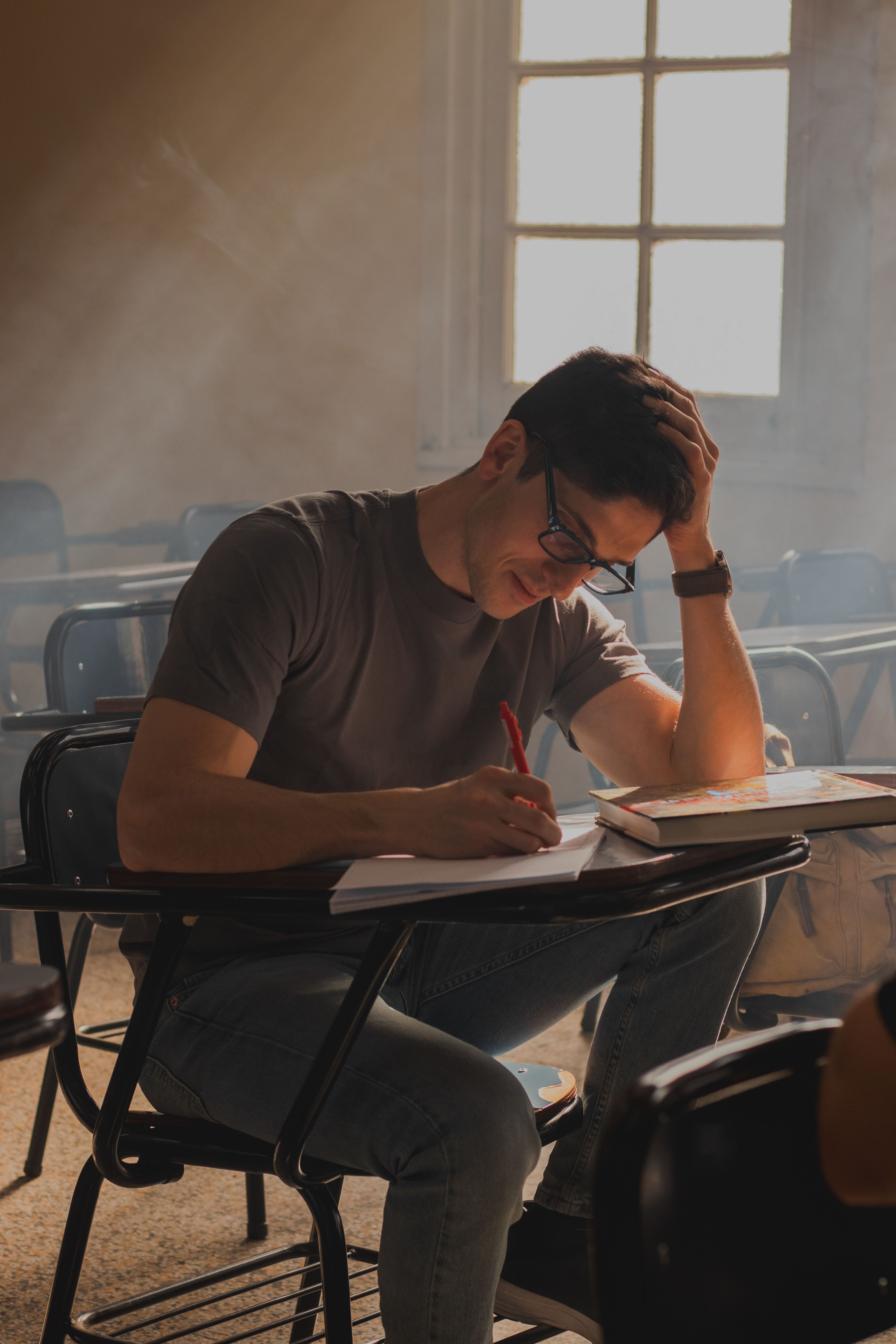 A concentrated young man studies in a sunlit classroom, surrounded by empty desks.