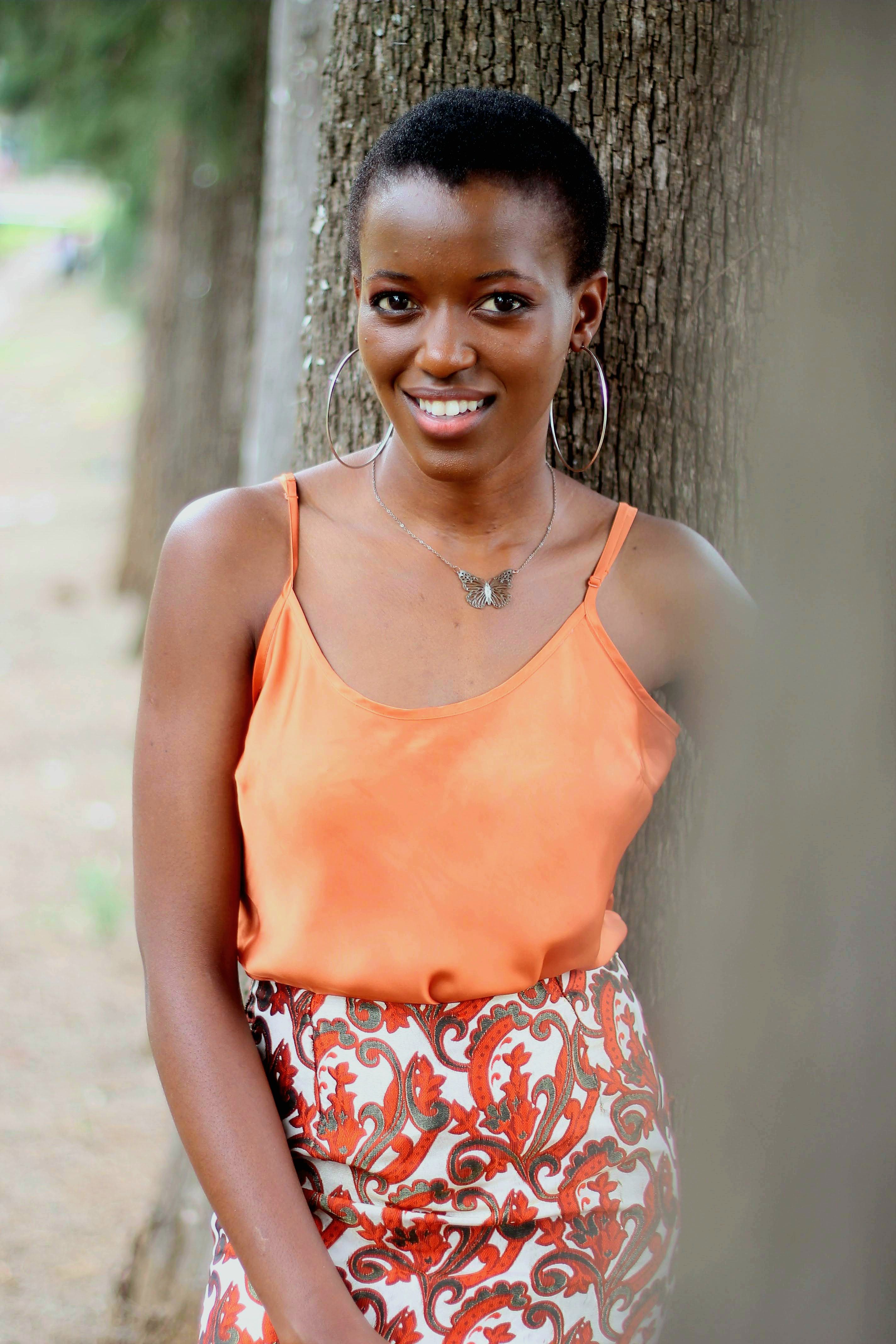 Smiling woman posing outdoors in an orange top with patterned skirt against a tree.