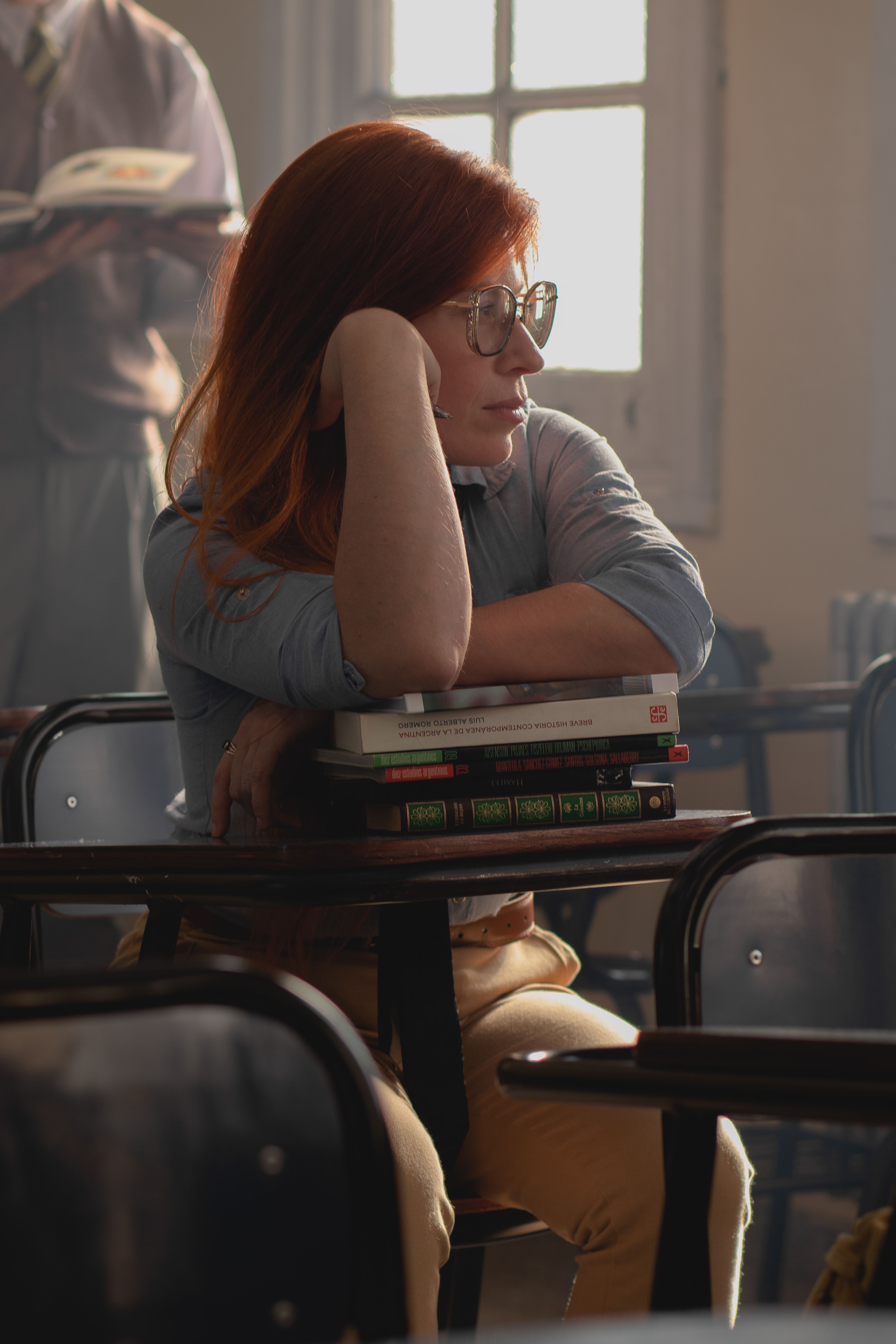 A thoughtful student with red hair leans on a desk full of books in a sunlit classroom.