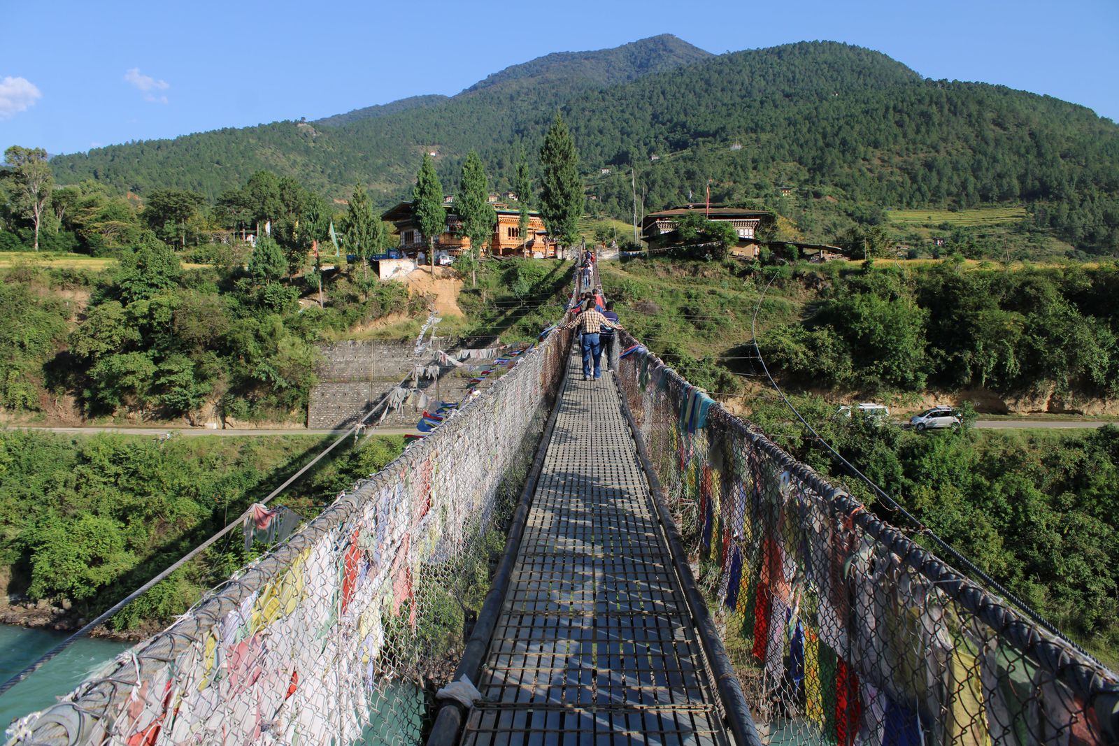 A long steel suspension footbridge hung with prayer flags over a green river in Bhutan, two walkers crossing to farmhouses