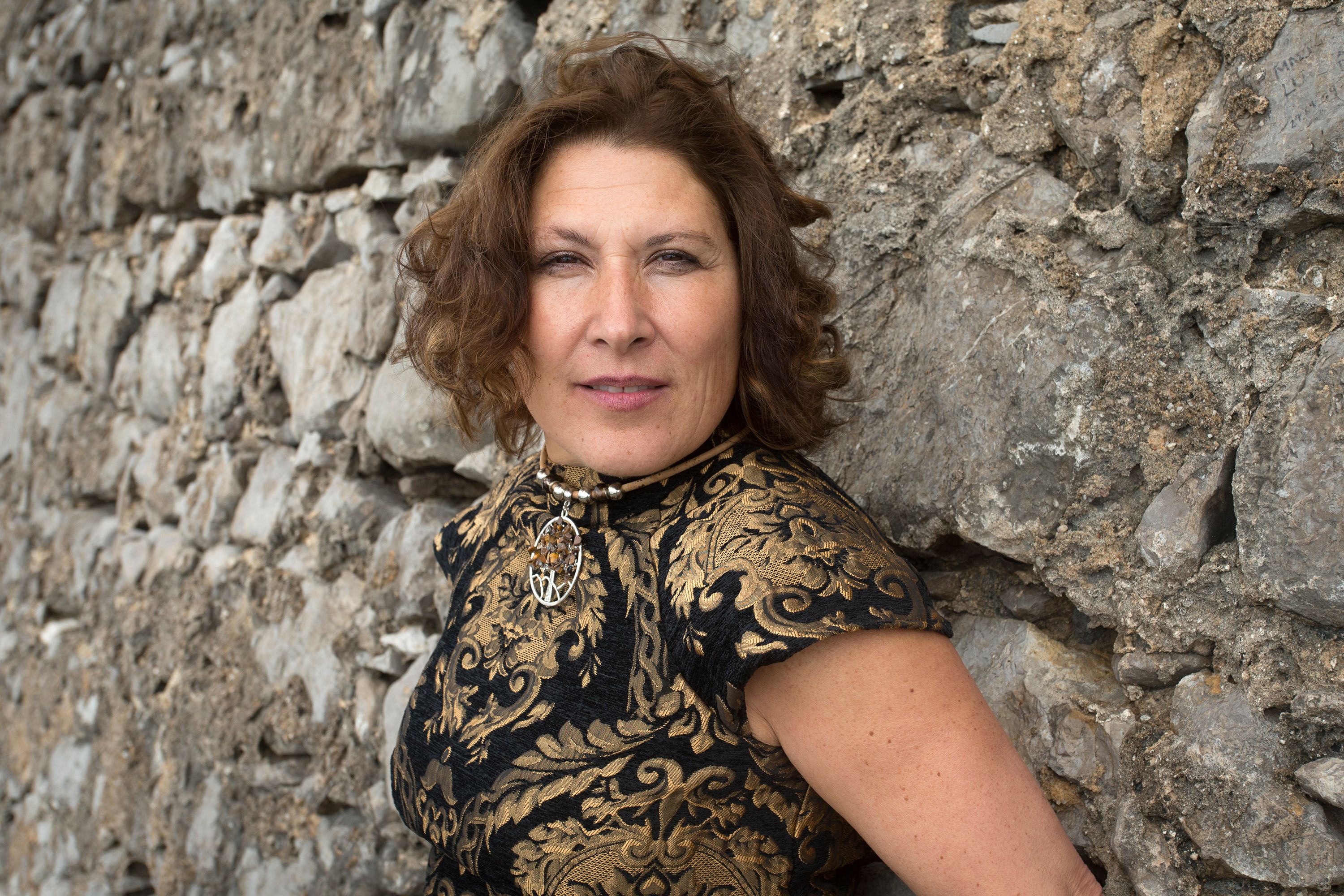 Elegant woman in patterned attire posing by a rustic stone wall.