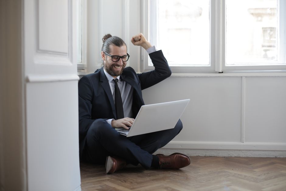 Homem alegre sentado no chão com um laptop, celebrando o sucesso no trabalho.