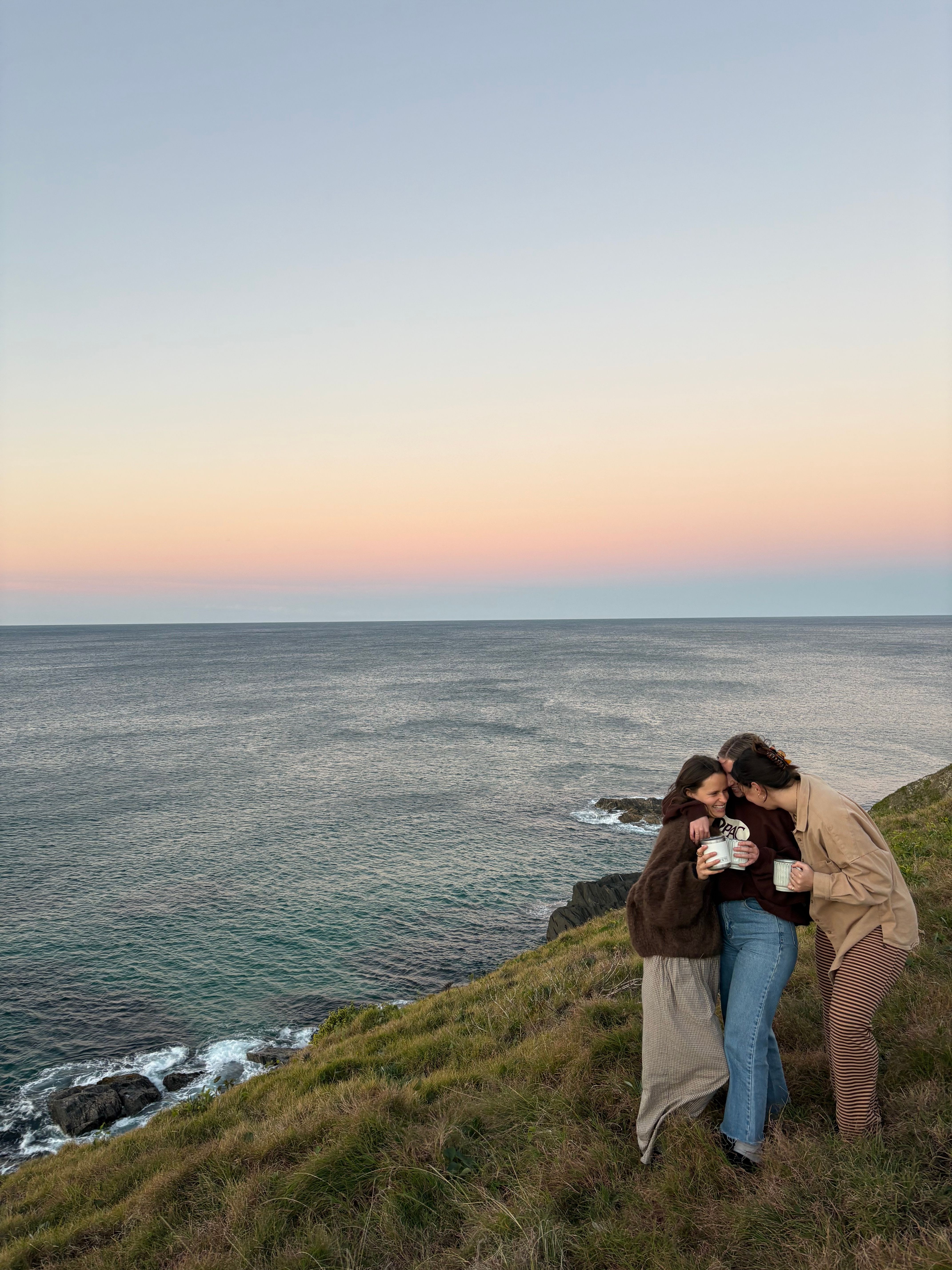 Three friends sharing a warm drink on a cliffside as the sun sets over the ocean in Sydney.