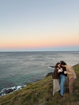Three friends sharing a warm drink on a cliffside as the sun sets over the ocean in Sydney.