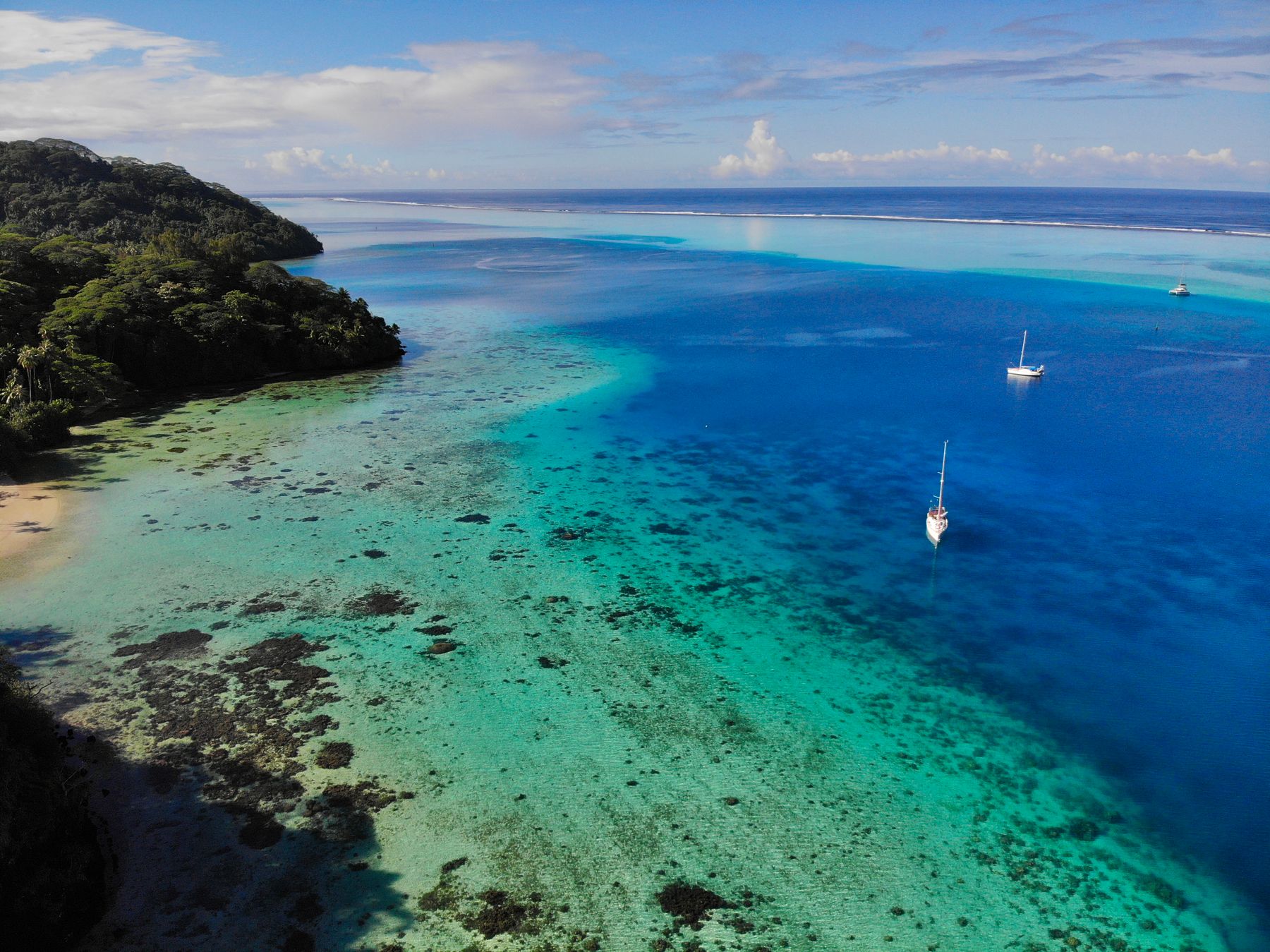 Sailboats moving across a reef-fringed French Polynesian lagoon between island chapters