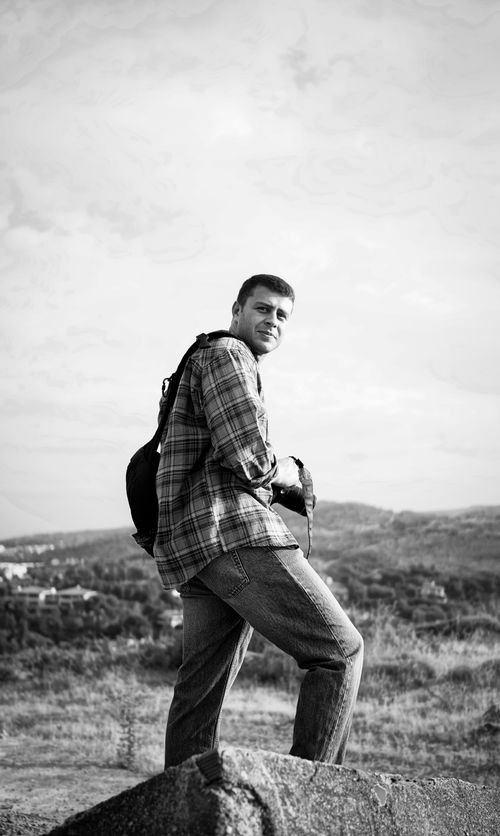 Free A man in casual attire holds a camera while hiking on a hilltop, showcasing a scenic landscape view. Stock Photo