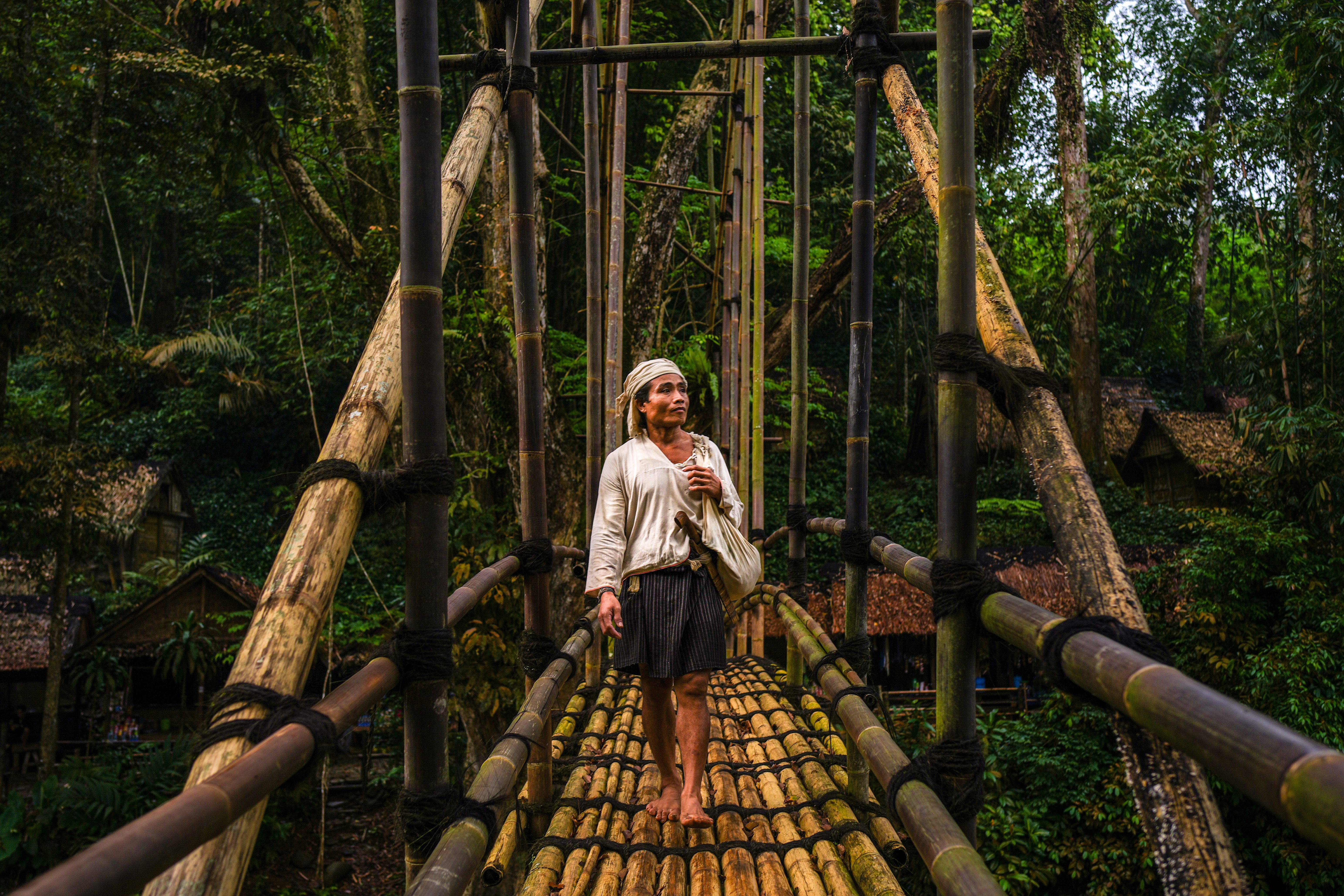 A person walks across a traditional bamboo bridge in a lush Indonesian jungle setting.