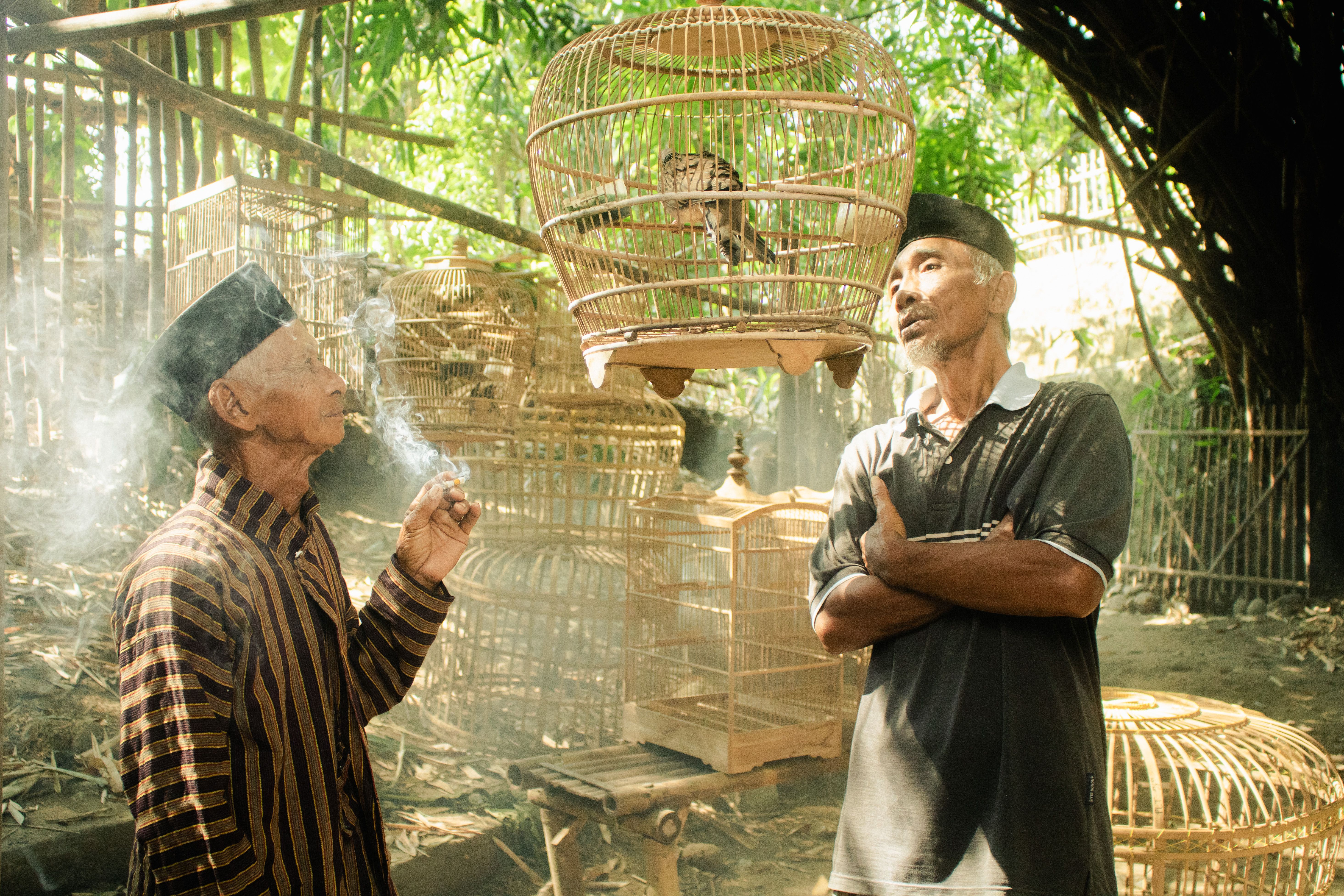 Two elderly men in traditional clothing stand outdoors, admiring a birdcage, exuding warmth and culture.