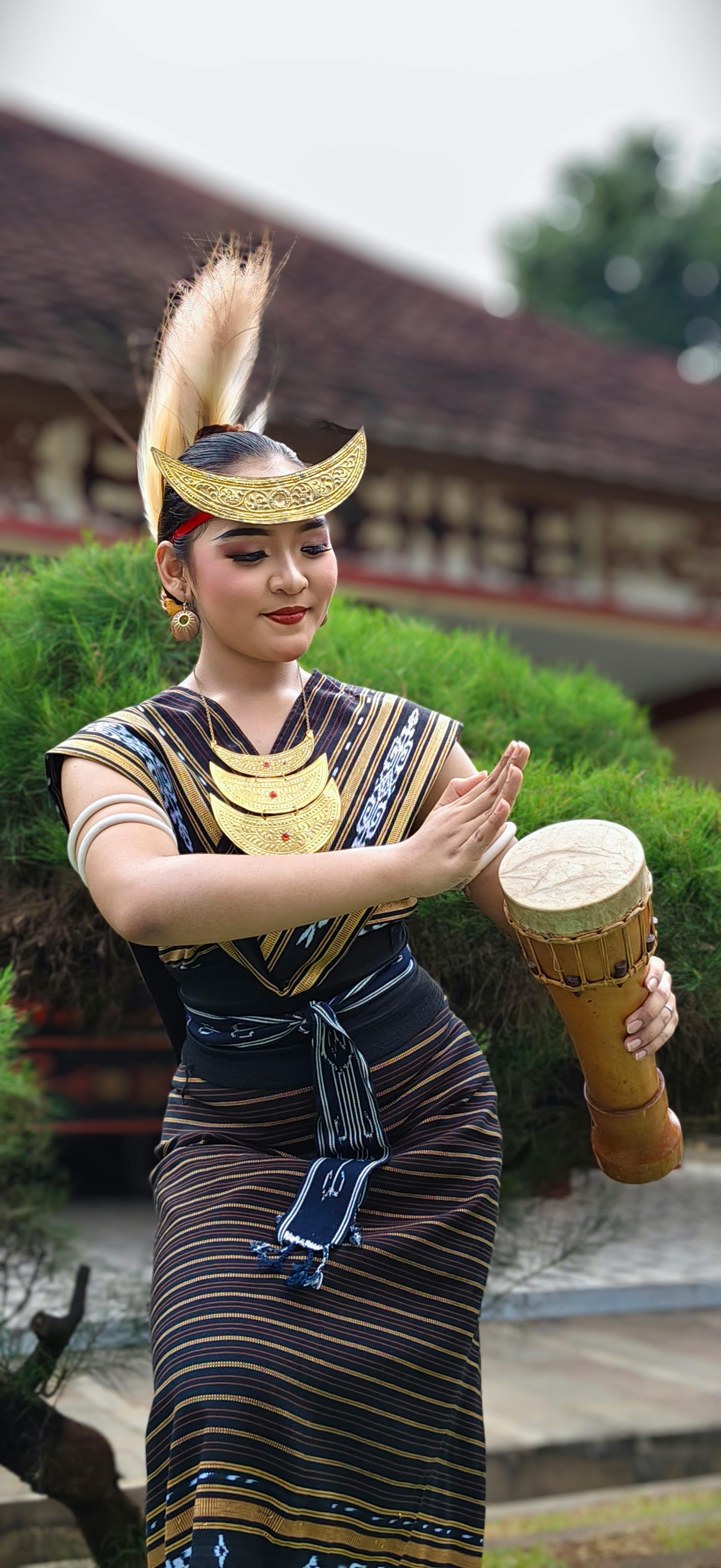 A woman in traditional attire performs a cultural dance while playing a drum outside.