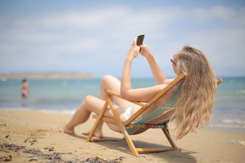 藍色和白色比基尼的女人坐在沙灘上的棕色木椅上，同時使用智能手機