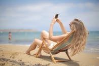 金发女子靠在椅子上，手里拿着智能手机，在海滩上享受阳光明媚的一天