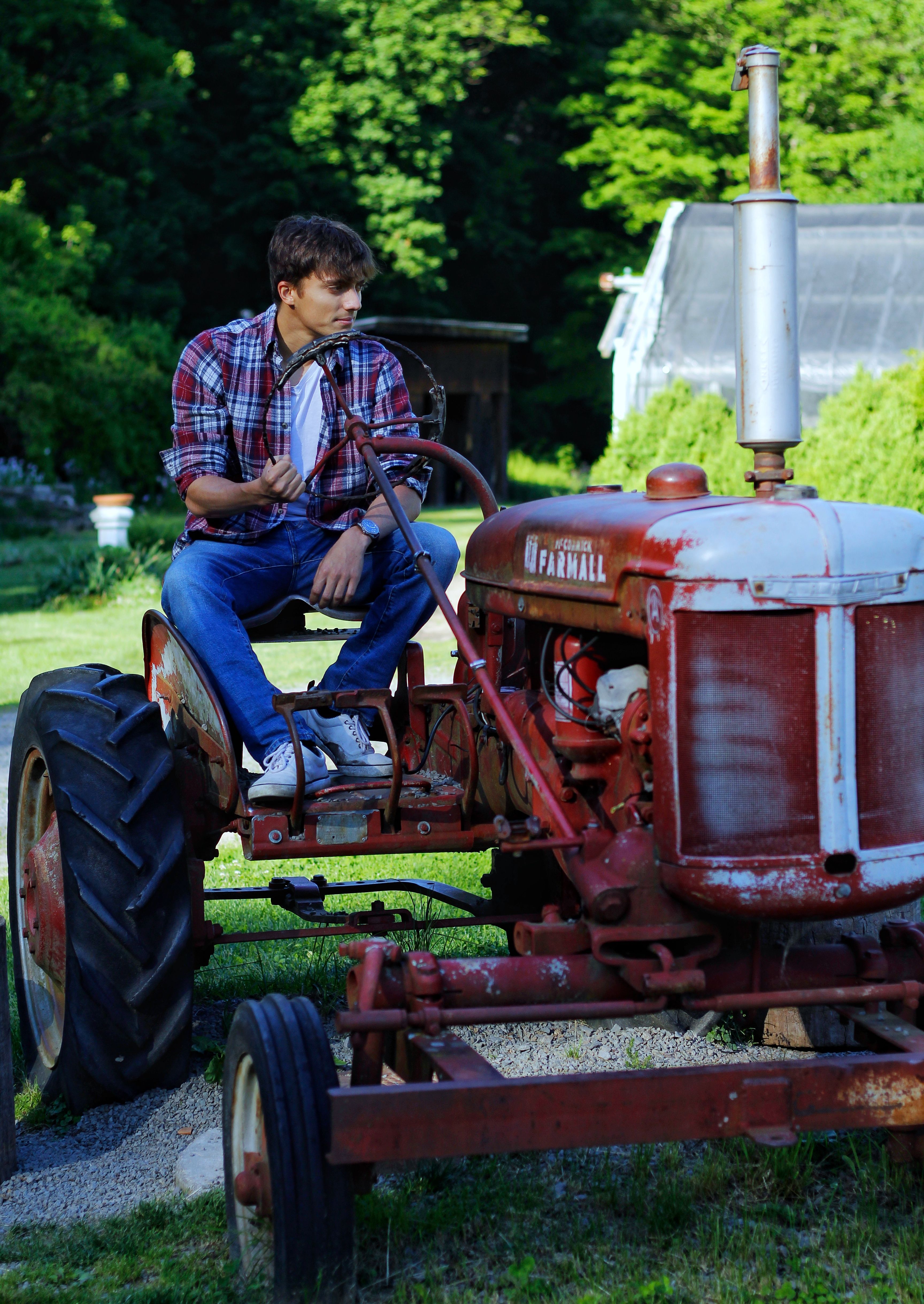 A young man sits on a vintage tractor surrounded by lush greenery.