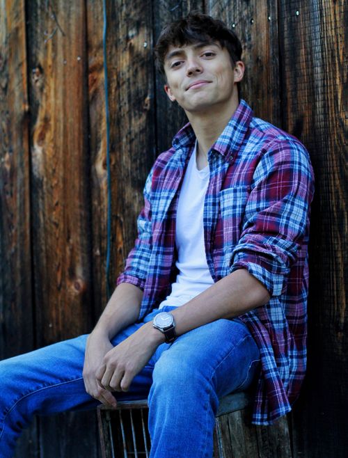 Free Young man wearing a plaid shirt sitting against a wooden wall outdoors. Stock Photo
