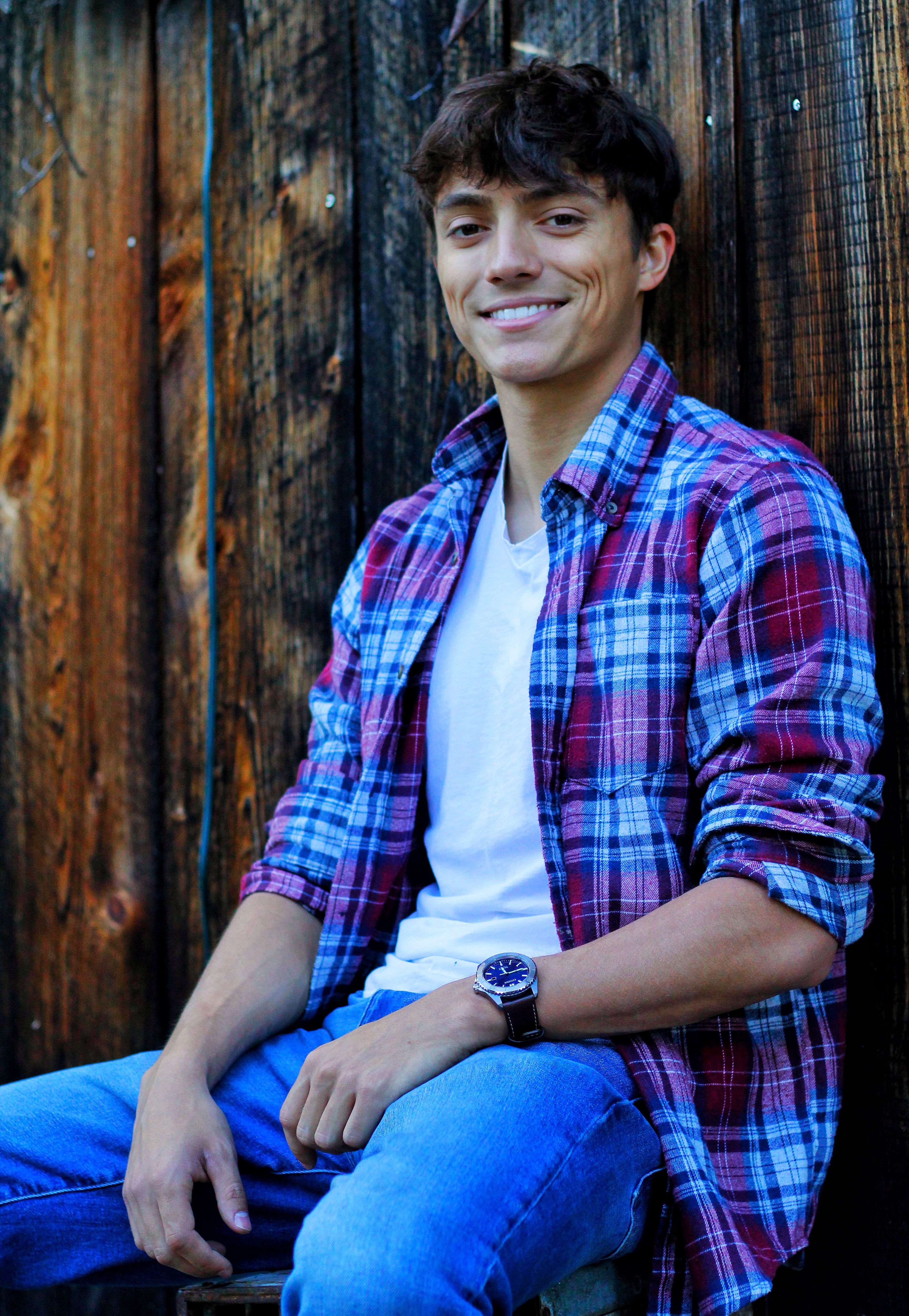 Young man smiling casually in checkered shirt and jeans against wooden backdrop.