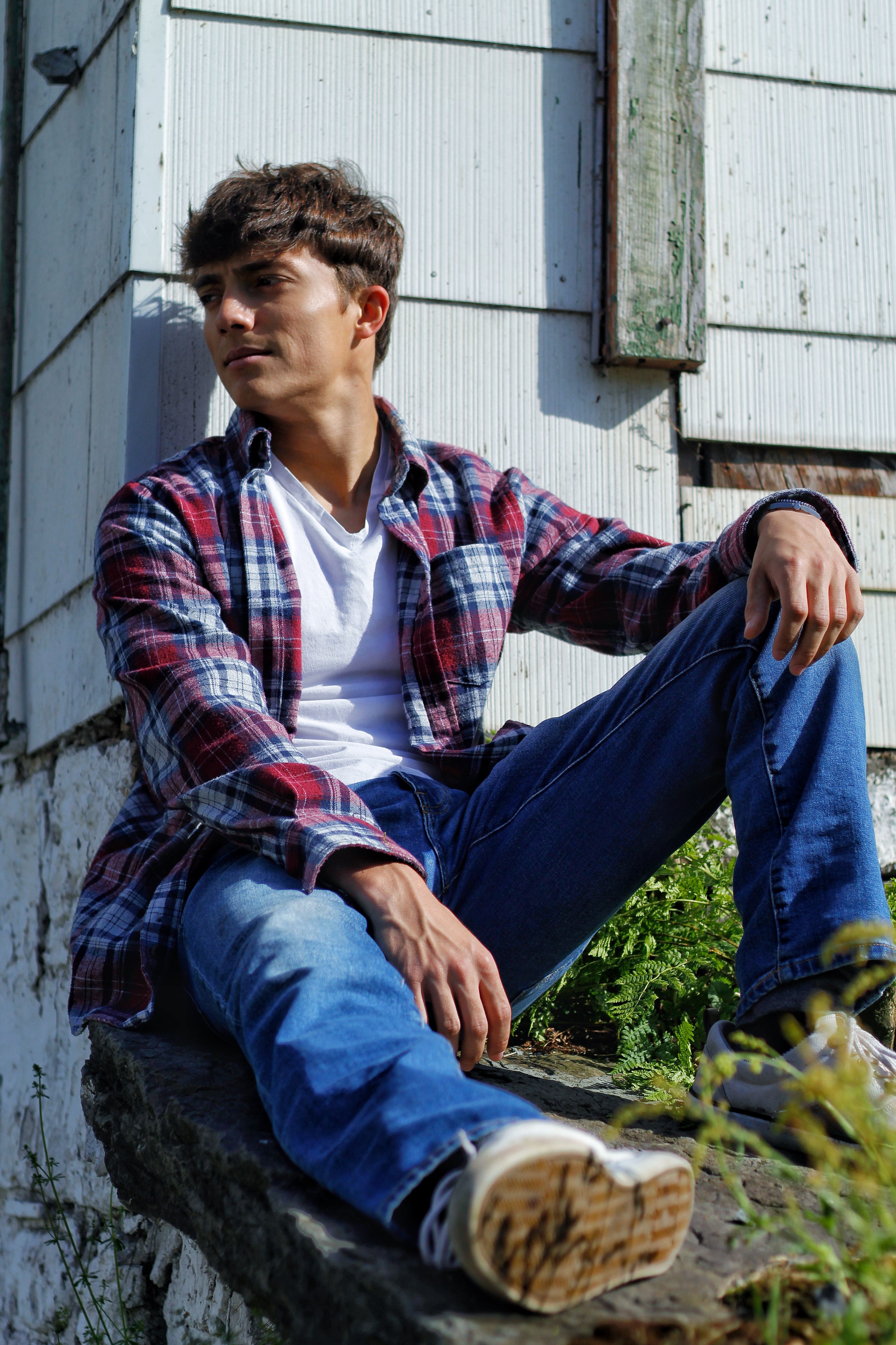 Casual young man sitting outside a rustic wooden house, enjoying a sunny day.