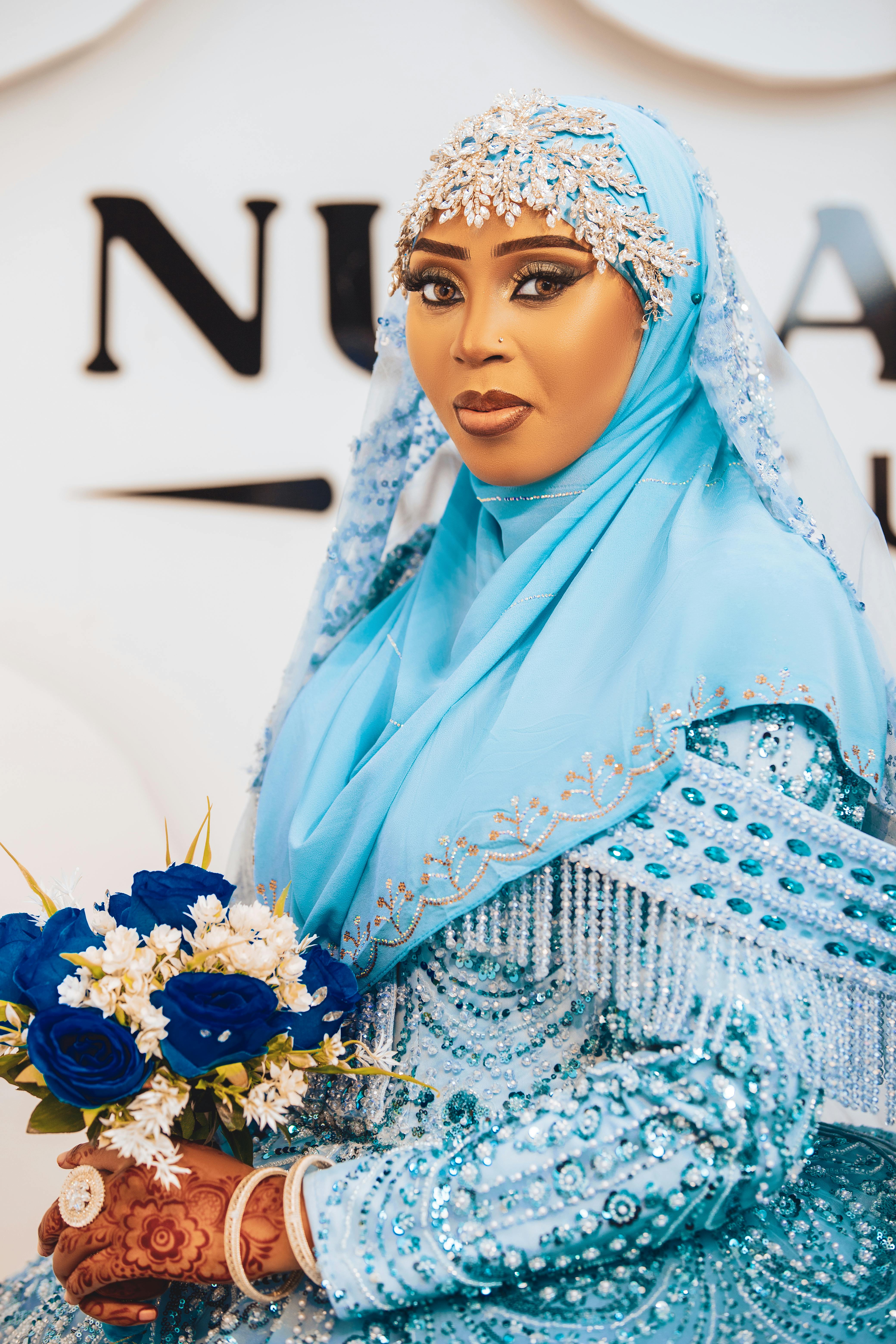 A bride in a beautifully detailed blue hijab holds a bouquet of blue and white flowers, exuding grace and elegance.