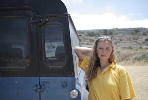 阳光明媚的乡村里，一名戴太阳镜的女子正倚着一辆蓝色汽车摆姿势