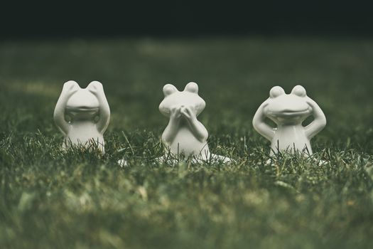 Three ceramic frog statues depicting the 'see no evil, hear no evil, speak no evil' proverb on grass.