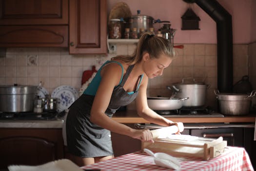A woman in an apron rolls dough on a kitchen counter, illuminated by warm natural light.