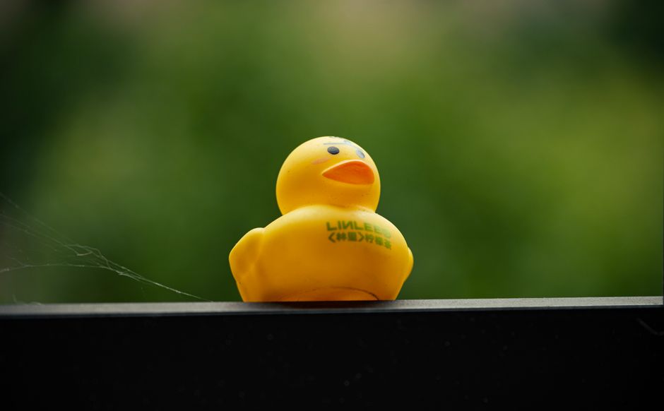 A yellow rubber duck sits on a balcony railing with a blurred green background, providing an outdoor playful vibe.