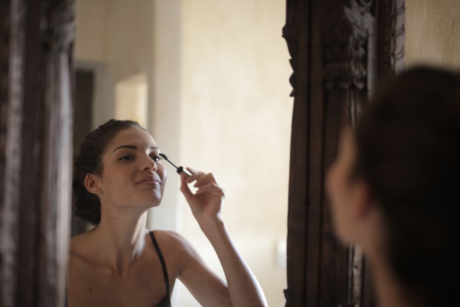 A woman applies mascara while looking in a stylish wooden mirror indoors.
