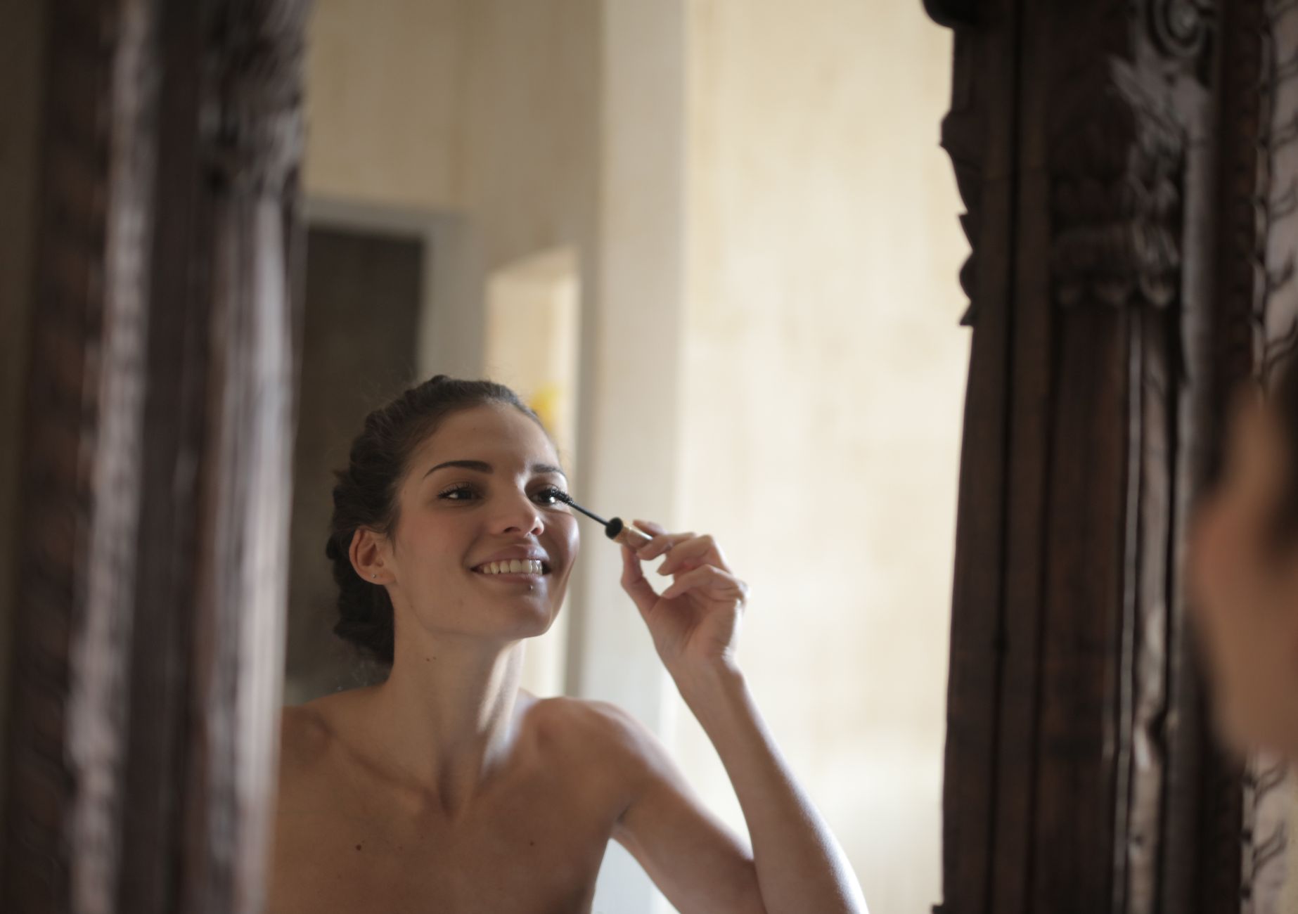 Smiling woman applying mascara in front of a vintage mirror.