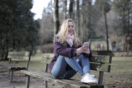 A woman sits joyfully on a wooden park bench during winter, texting on her smartphone.