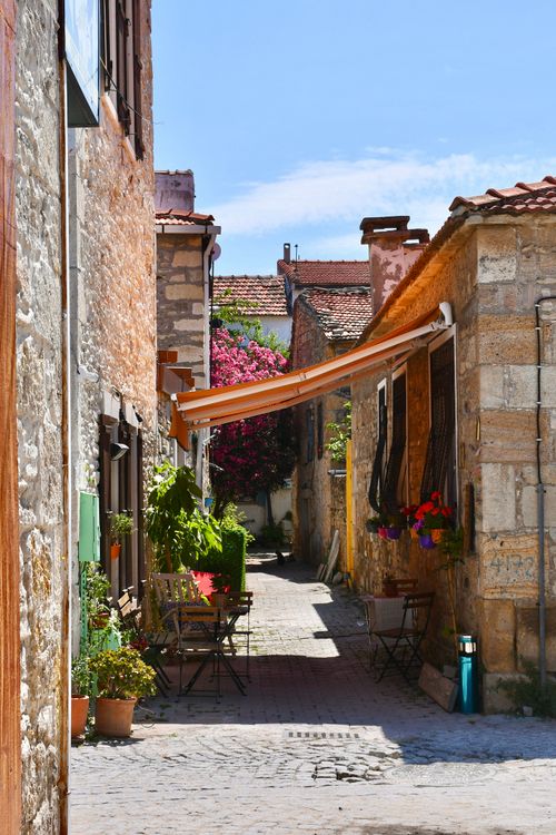 Free A picturesque Mediterranean alley featuring stone buildings, flowering plants, and an orange sunshade in bright daylight. Stock Photo