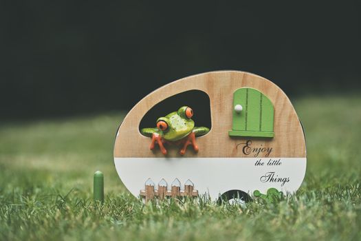 A cute frog toy peeks through a decorative wooden caravan sign with 'Enjoy the little things' text.