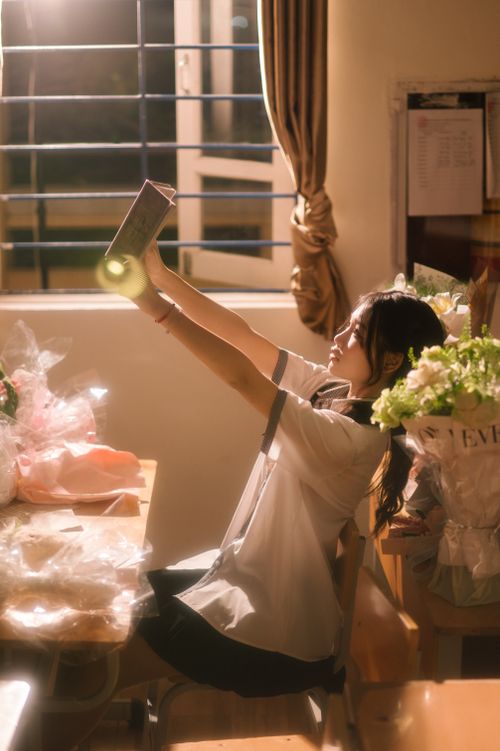 Free A young woman enjoys reading indoors near a window, surrounded by flowers and soft light. Stock Photo
