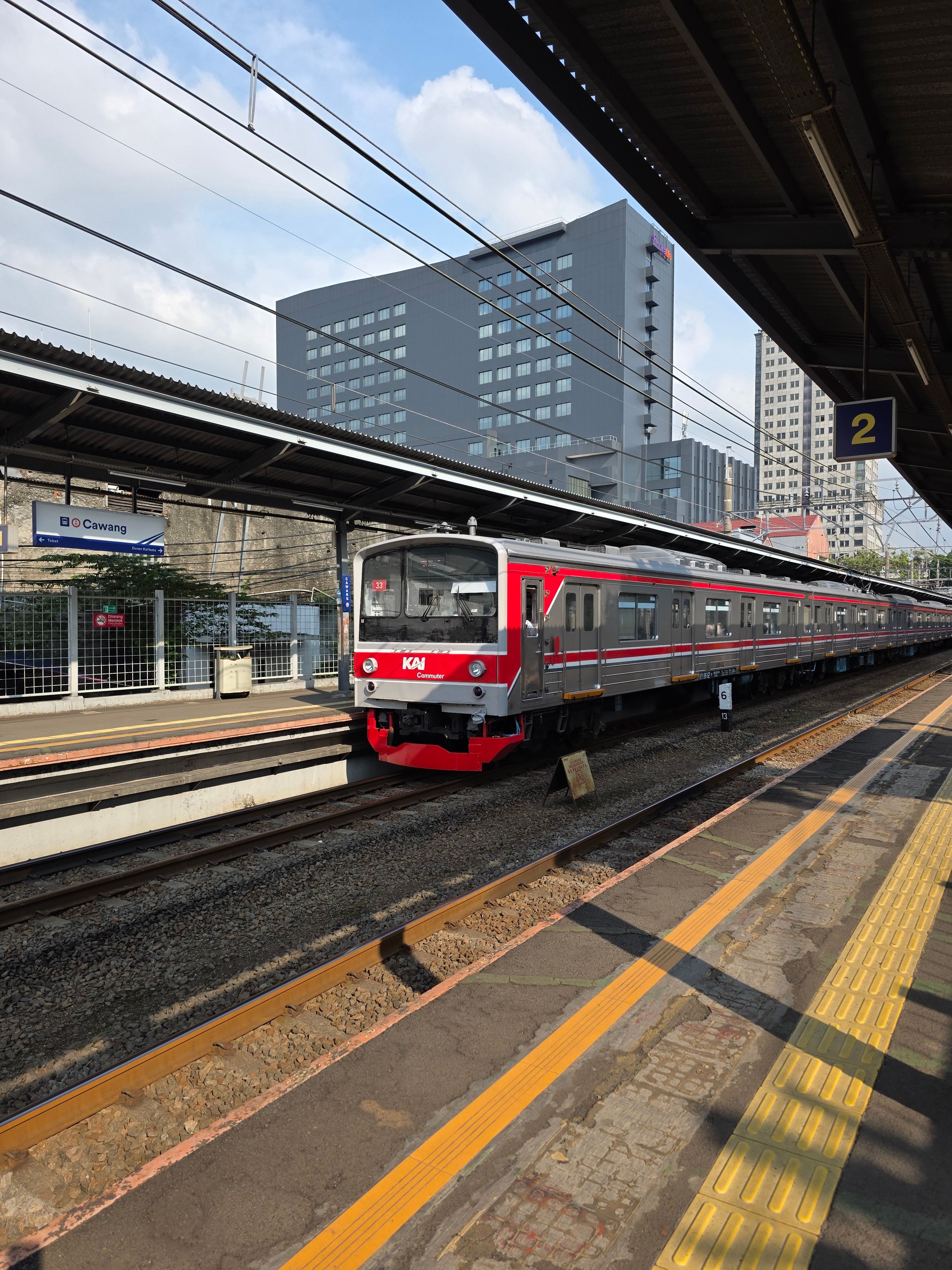 Red commuter train arriving at a Jakarta station during the day under a blue sky.