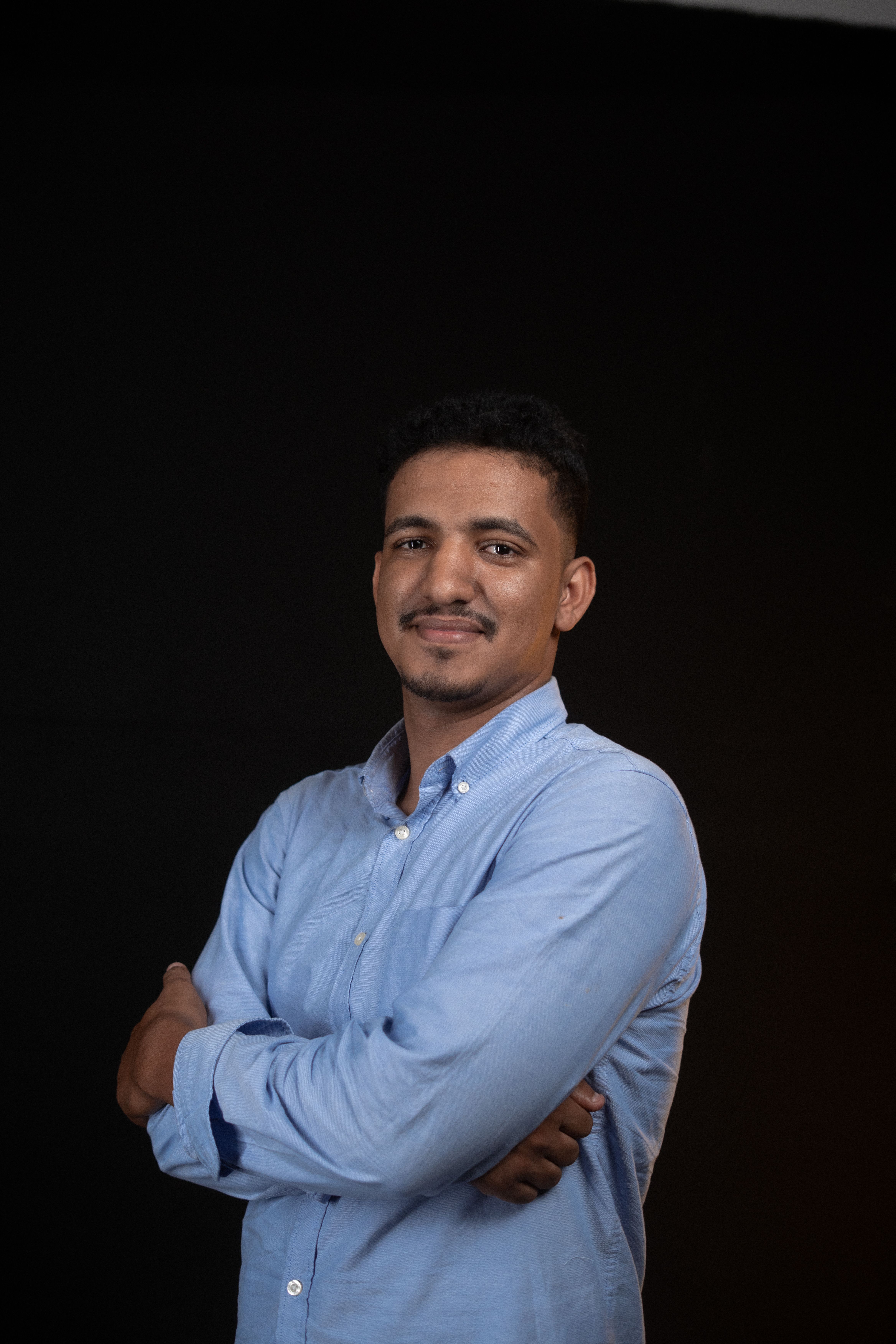 Portrait of a smiling man in a blue shirt with arms crossed, studio setting.