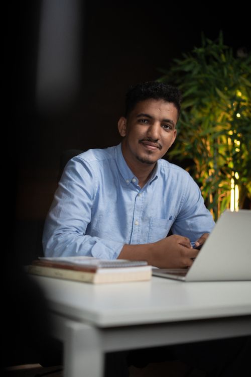 Free Young man working on a laptop at home, sitting at a desk with books. Stock Photo