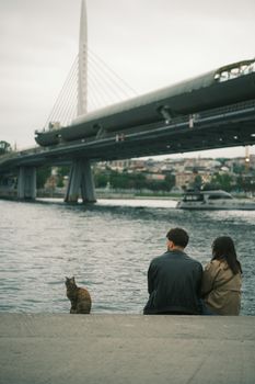A couple and a cat sit by the water with a modern bridge in the background. Overcast city scene.