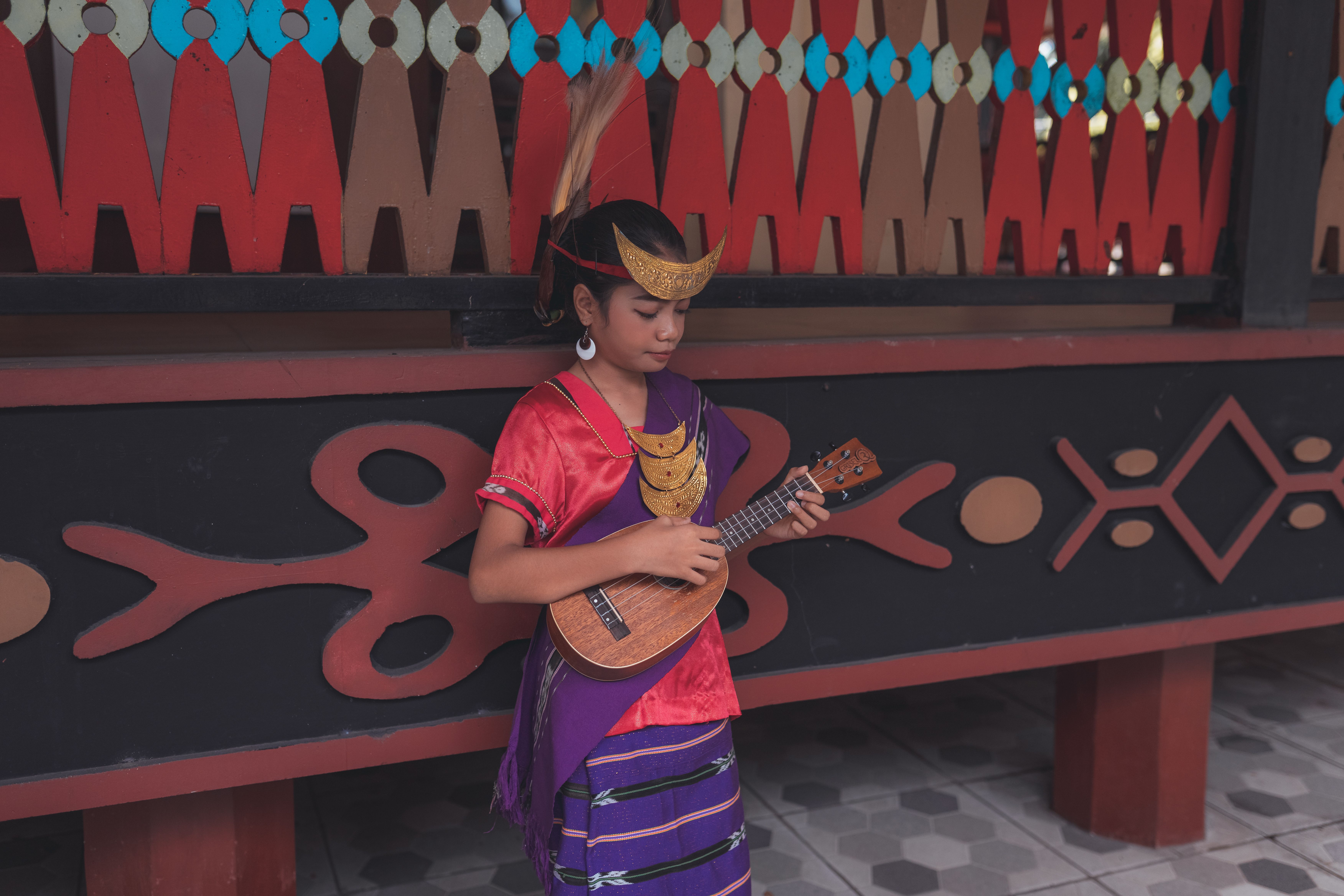 Young girl in traditional attire playing ukulele against cultural backdrop in Jakarta.