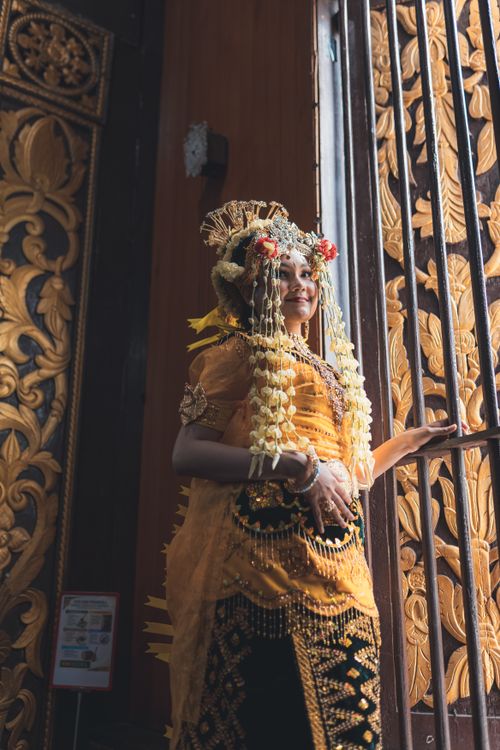 Free Beautiful Balinese bride in traditional attire stands by ornately carved wooden door. Stock Photo