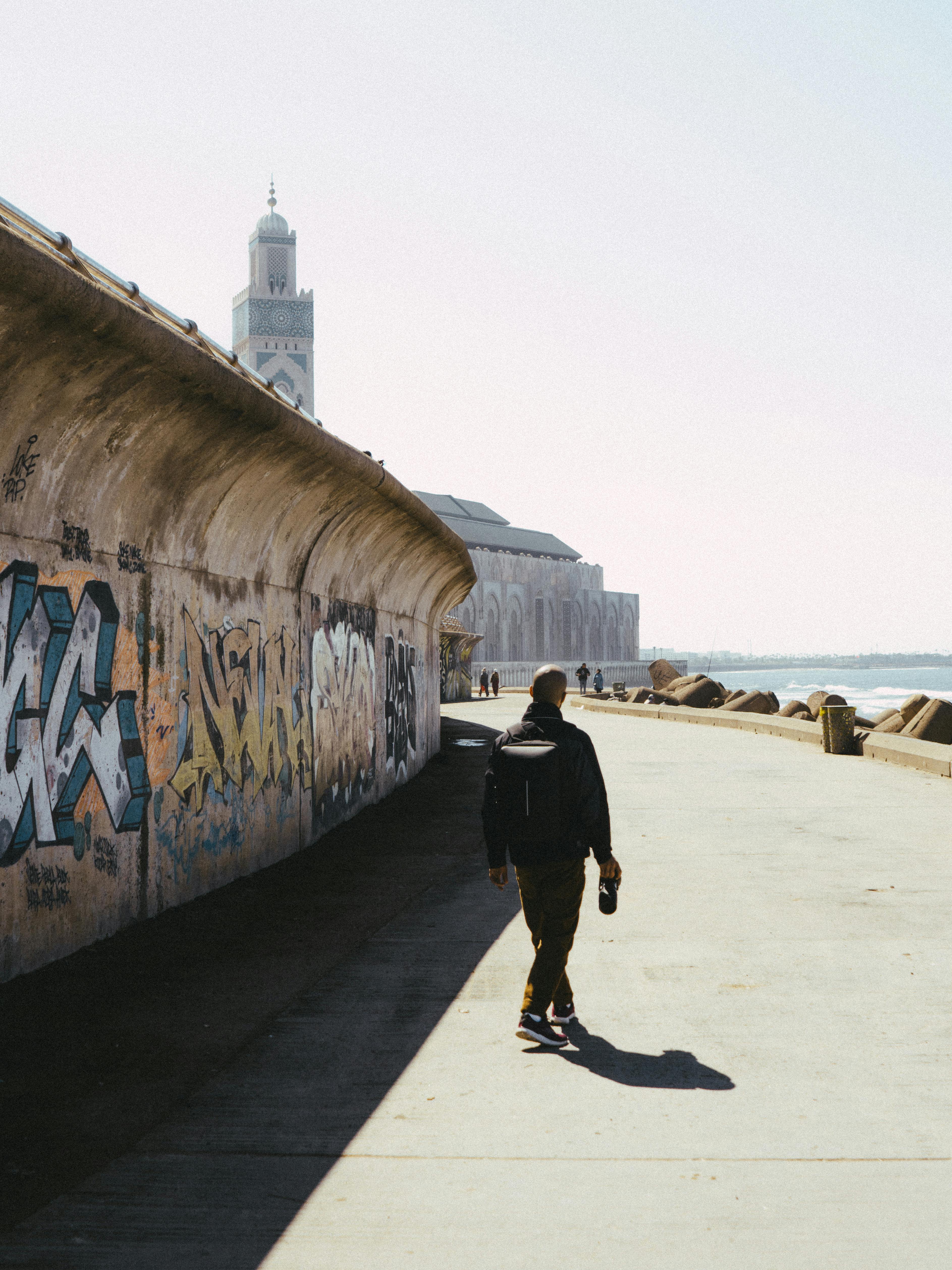 Person walking along graffiti wall next to Hassan II Mosque during daytime in Casablanca, Morocco.