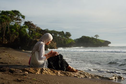 A woman in a hijab reads by the beach in Jawa Barat, Indonesia during a serene day.