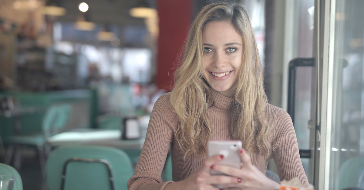 A person holding a basic flip phone in a coffee shop, looking relaxed and engaged with their surroundings