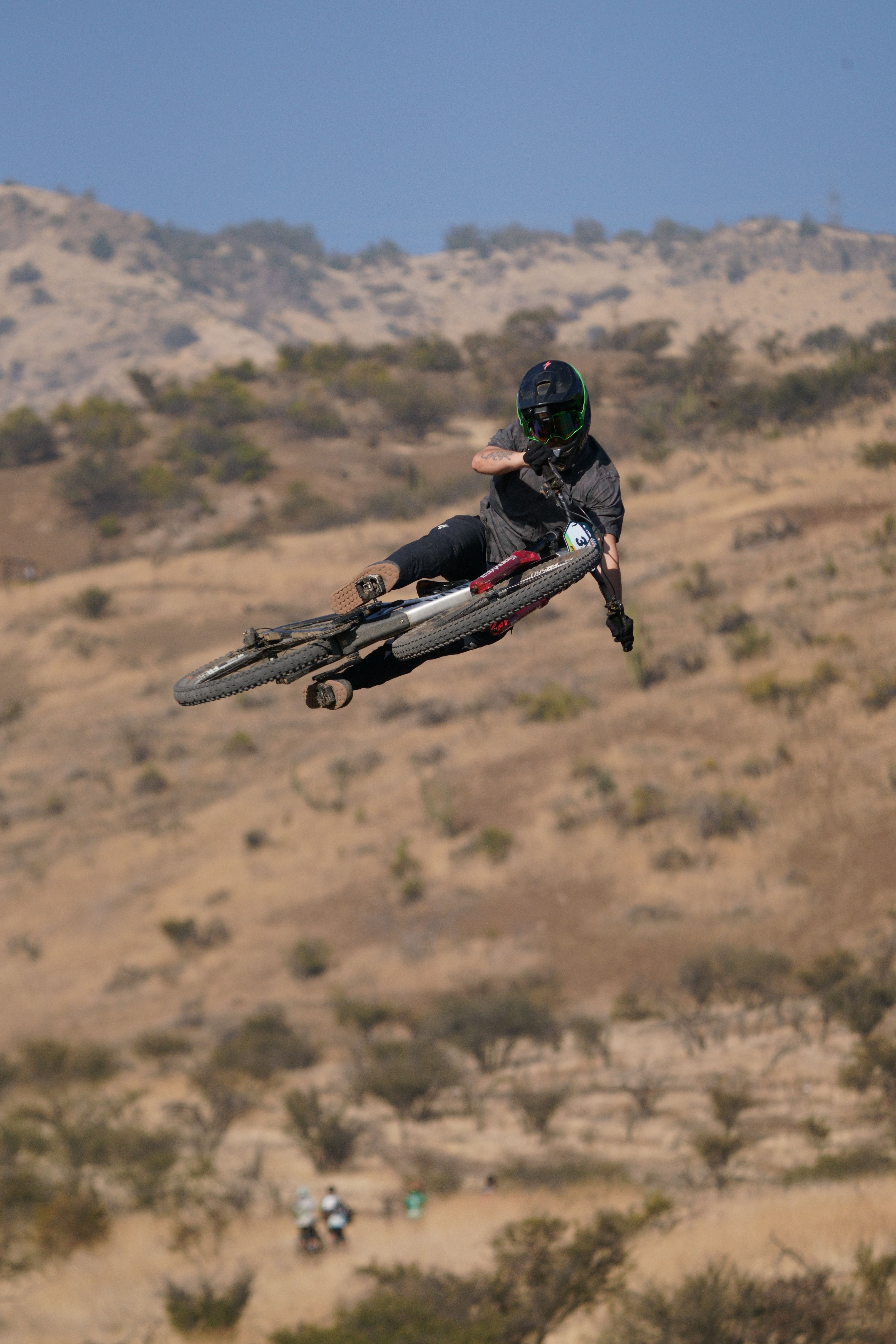 Rider performing an impressive jump on a mountain bike in a dry, hilly landscape.