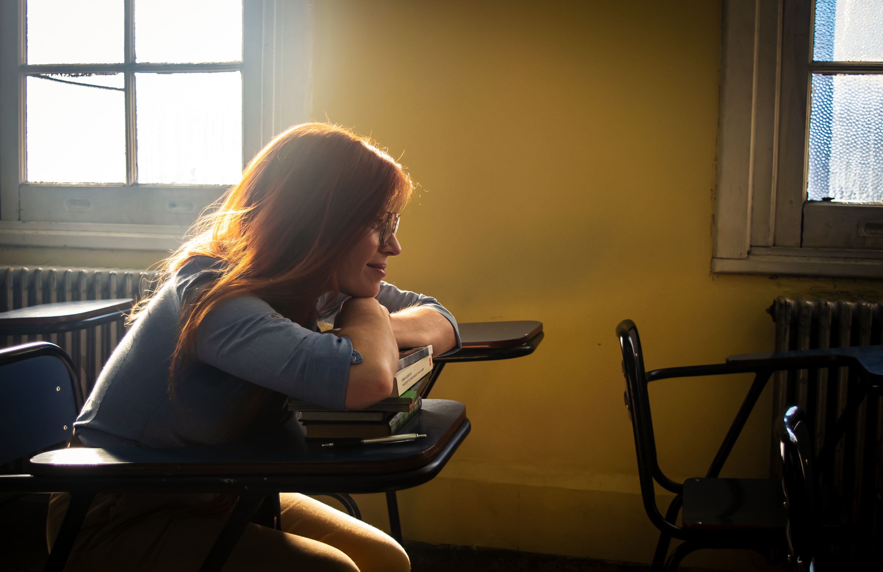 A thoughtful young woman with red hair sitting in a sunlit classroom, leaning on a desk with a dreamy expression.