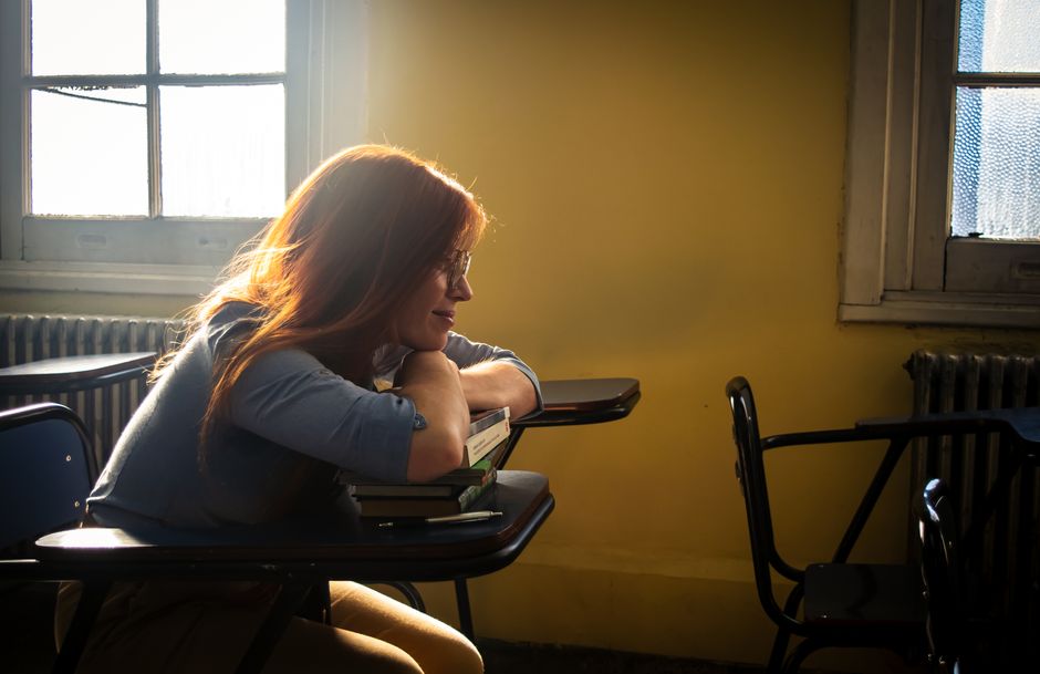 A thoughtful young woman with red hair sitting in a sunlit classroom, leaning on a desk with a dreamy expression.